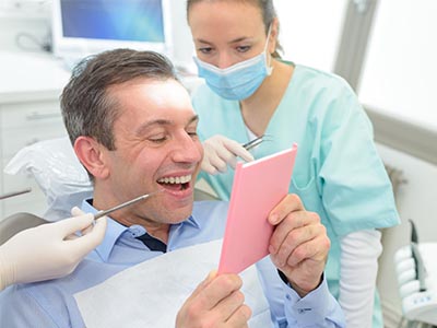 A man in a dental chair, holding up a pink card with a surprised expression, while a dentist and hygienist look on.