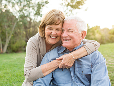 An elderly couple embracing in a park, with the man wearing a blue shirt and the woman in a white top.