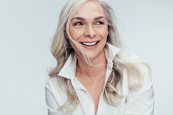 The image shows a woman with short, blonde hair smiling at the camera. She has a light complexion and is wearing a white top. Her expression appears friendly and approachable.