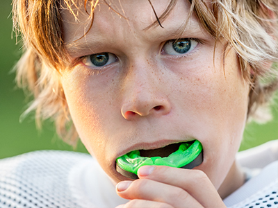 A young male with blonde hair and a sports jersey is brushing his teeth, holding a toothbrush in one hand and a green toothpaste tube in the other.