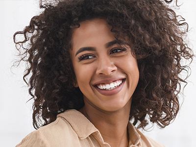 The image shows a woman with curly hair, smiling and looking directly at the camera. She is wearing a dark top and has a radiant complexion.