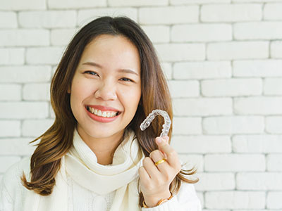 The image shows a woman smiling at the camera, holding up a clear plastic object that appears to be a dental appliance or mouthguard, with a brick wall in the background.